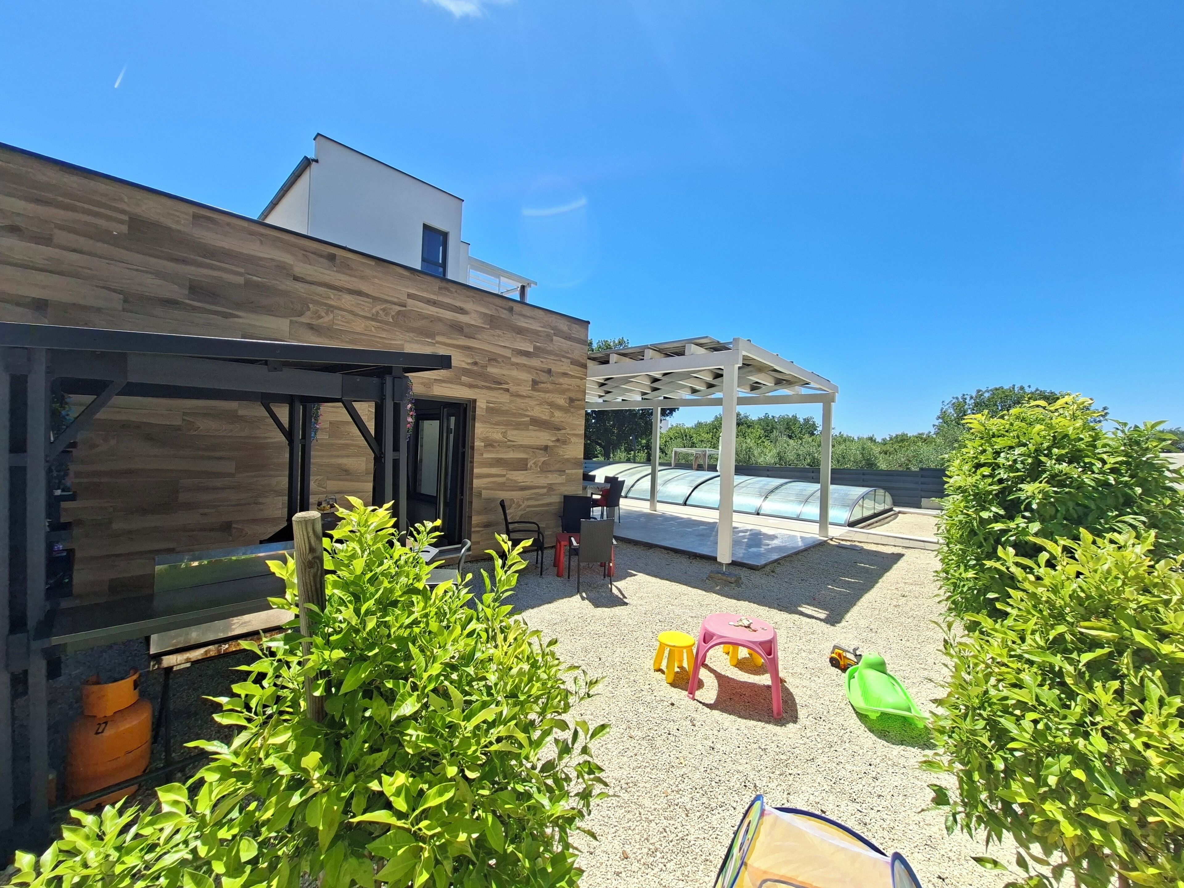 A modern outdoor patio with a wooden wall, a covered seating area, and a greenhouse structure.