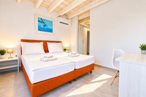 Ein Doppelbett mit weißen Bettlaken und einem orangefarbenen Kopfteil, ein Nachttisch mit Lampe sowie ein Topf mit Pflanze.