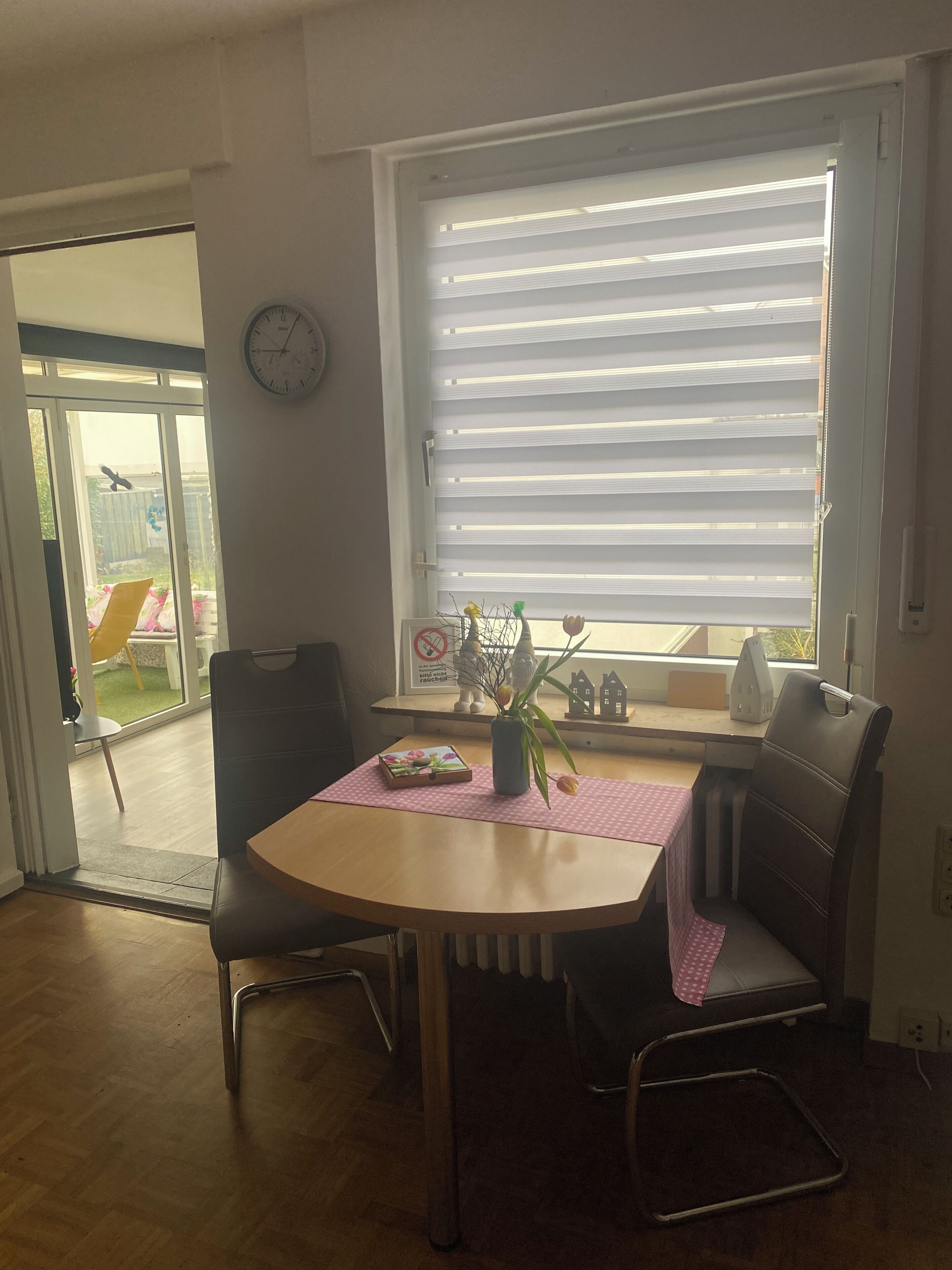 Ein Essbereich mit Tisch, Stühlen, einer Uhr und einem Fenster mit Jalousien.