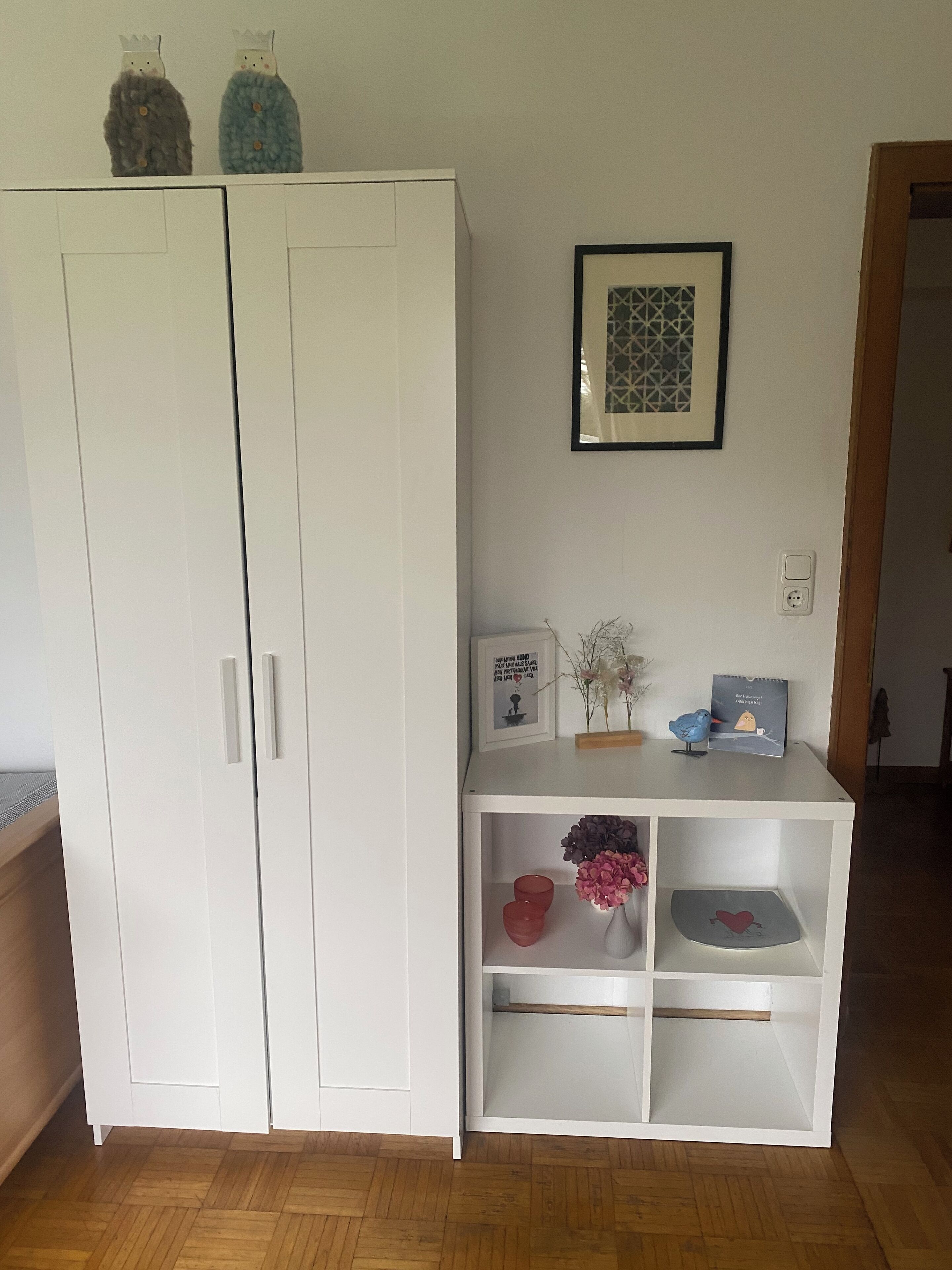 Un armario blanco con dos puertas, un estante blanco decorado con objetos y un cuadro colgado en la pared.