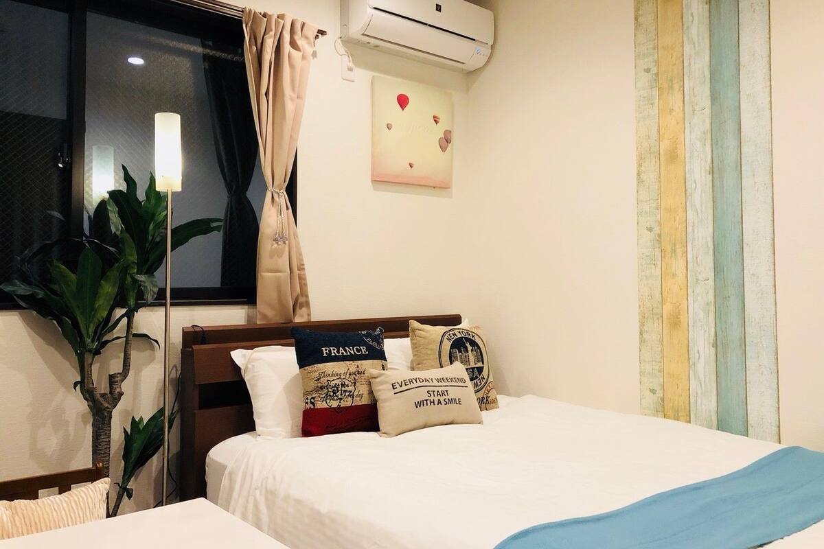 A hotel room with a bed, a nightstand, a potted plant, an air conditioning unit, and a window with curtains.