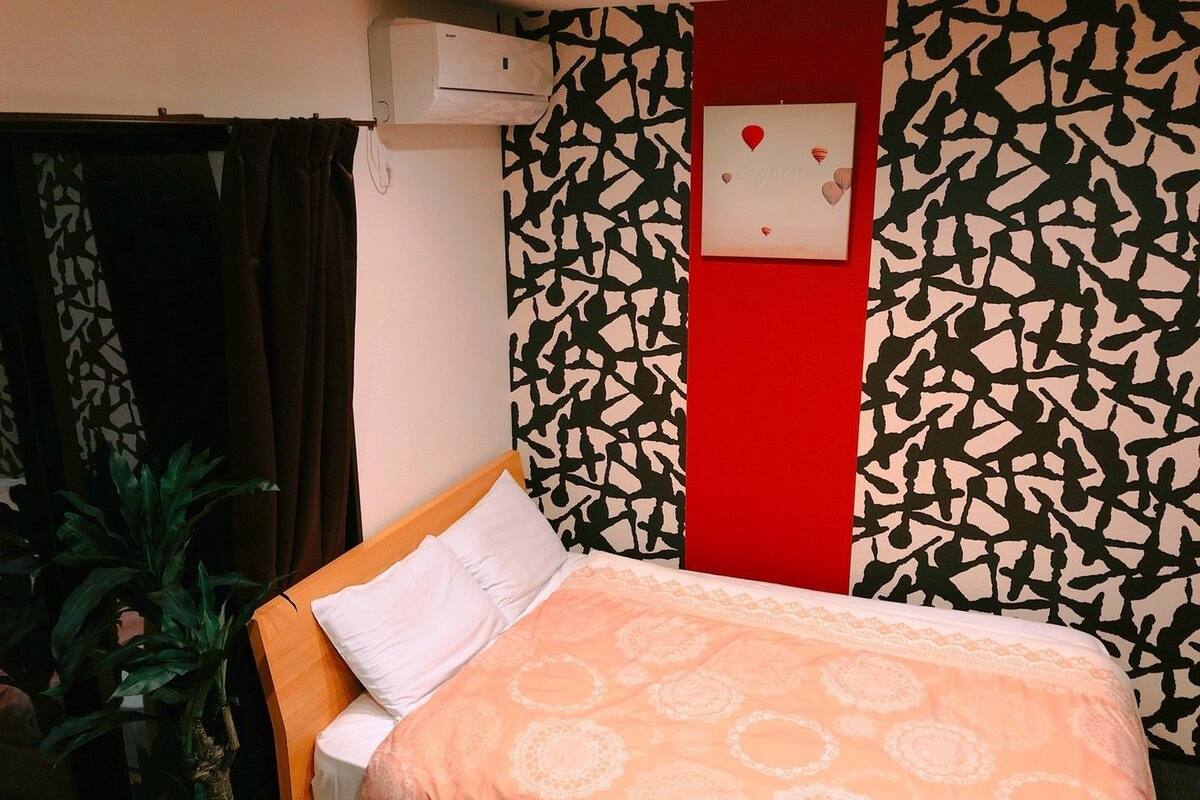 A hotel room with a bed, a potted plant, a red accent wall, and a patterned wall.
