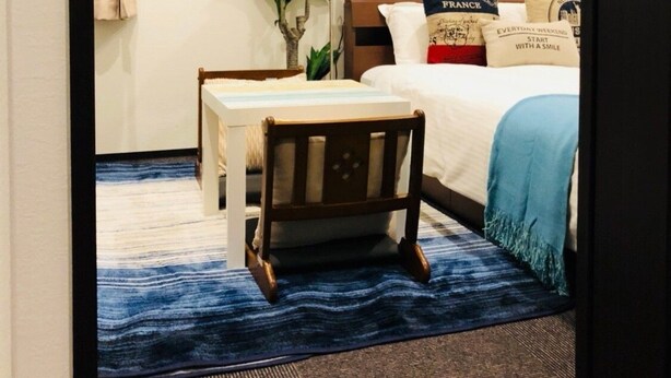 Quarto de hotel com cama, escrivaninha e cadeira. Há uma janela com cortinas, um vaso de planta e um ar condicionado fixado na parede.