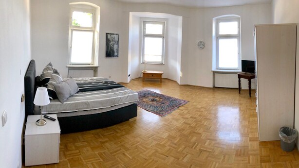 A bedroom with a bed, bedside table, lamp, and a small wooden cabinet. There are two windows, a radiator, and a door.