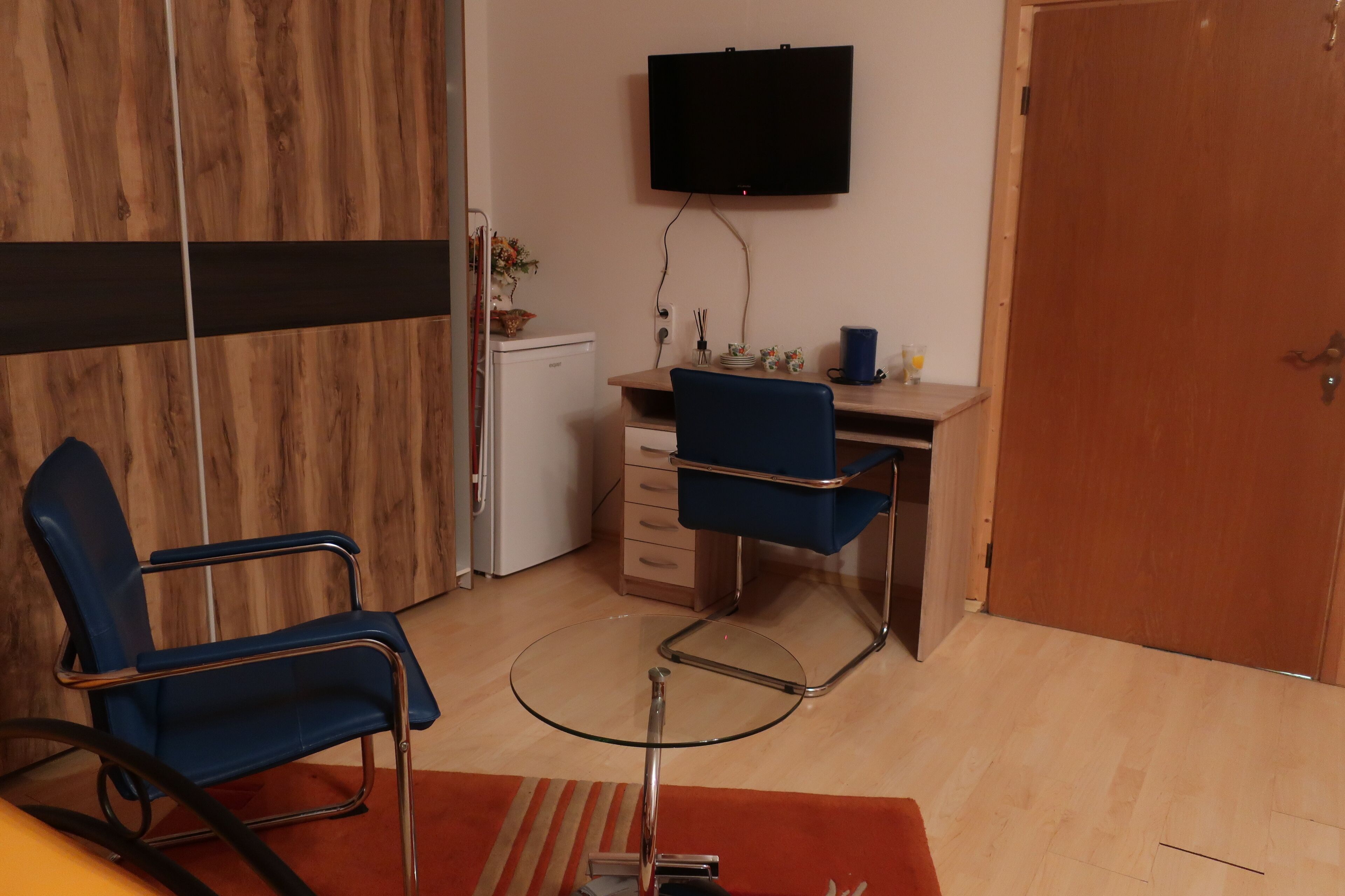 A room with a wooden wardrobe, a desk with a chair, a television mounted on the wall, and a small refrigerator.