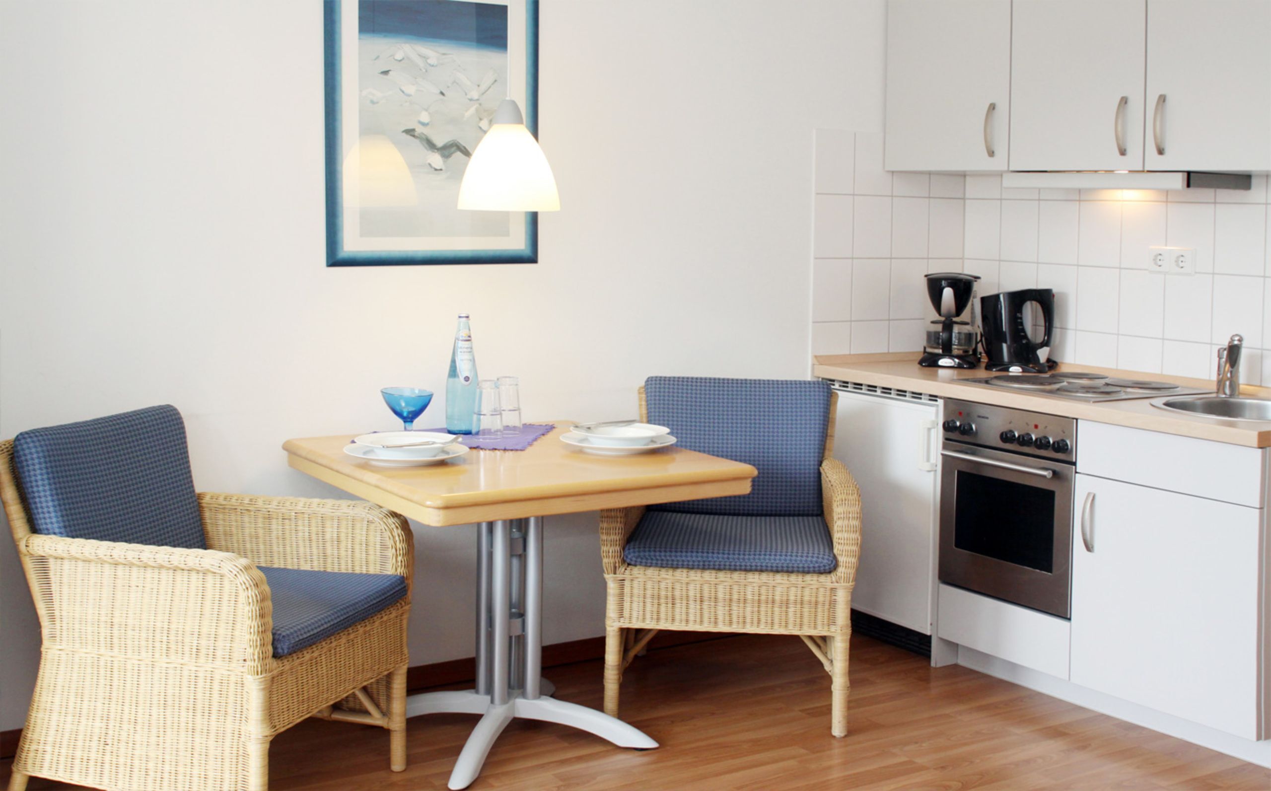 Une petite cuisine avec des meubles blancs, une cuisinière et un évier. On y trouve une table avec des chaises et un tableau encadré accroché au mur.