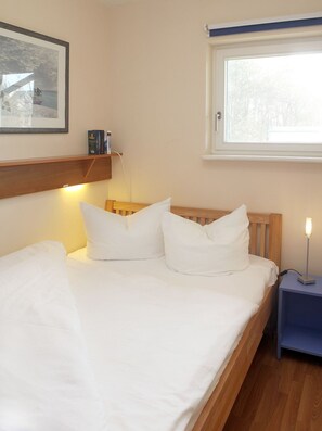 A neatly made bed with white linens and pillows, a bedside table with a lamp, a framed picture on the wall, and a window with a view of trees.