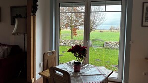 A dining area with a table set for two, a potted plant, and a view of a green landscape through a glass door. - House "NordSeeStern", detached, right on the North Sea dike with a fireplace (Wurster Nordseeküste)