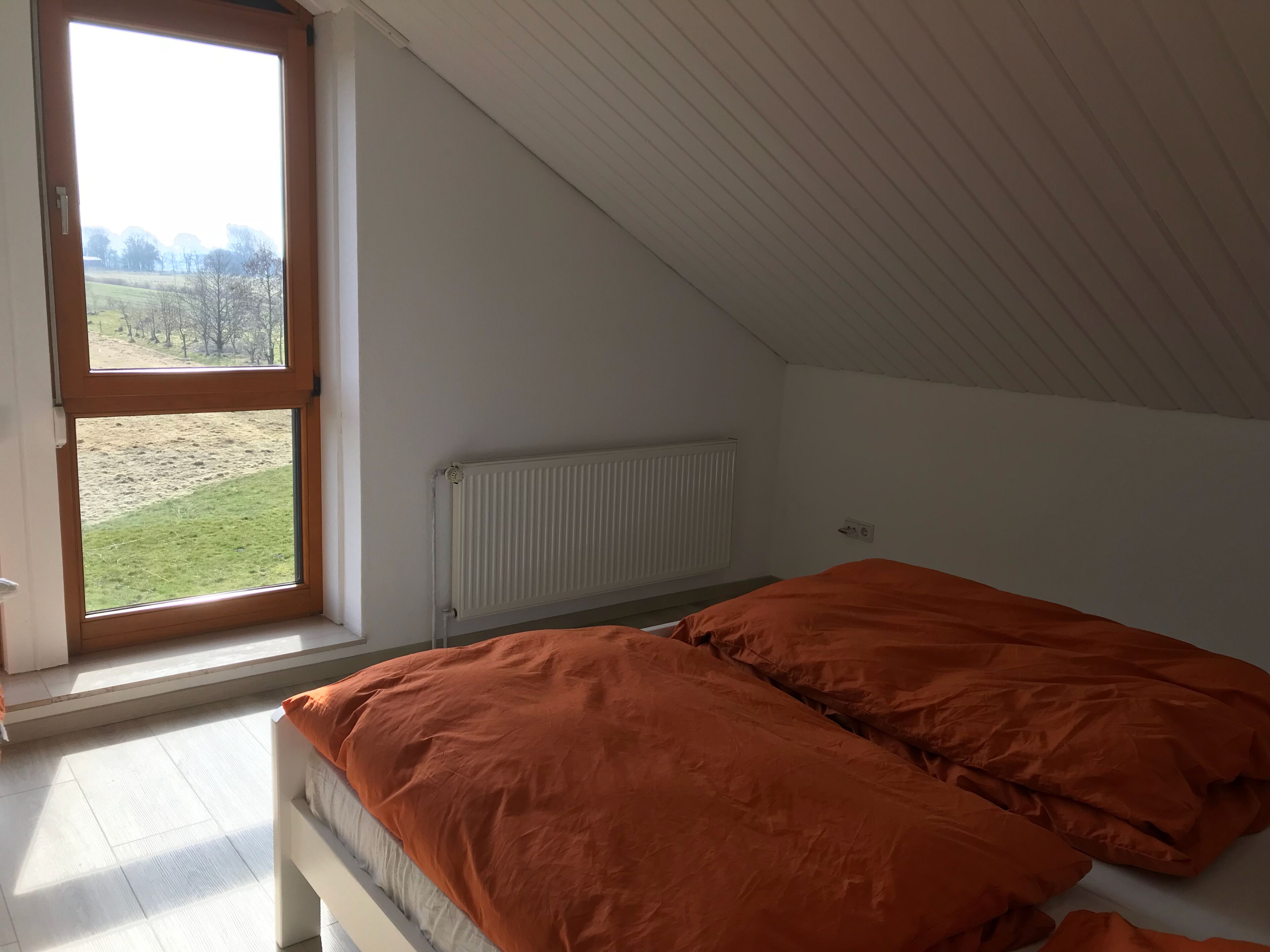 A bedroom with a large window, a white radiator, and a bed with orange bedding.