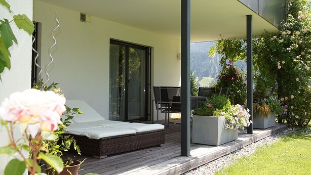 Un área de estar exterior moderna con un sofá, una mesa de comedor y sillas. Hay plantas en macetas y una vista de las montañas al fondo.