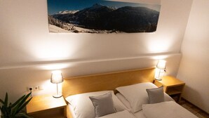 Cama doble con dos almohadas, mesita de noche con lámpara y un cuadro colgado en la pared que muestra un paisaje de montaña.