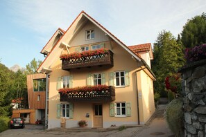 Un bâtiment à deux étages doté d’un balcon, une voiture garée devant et un mur de pierre sur la droite.