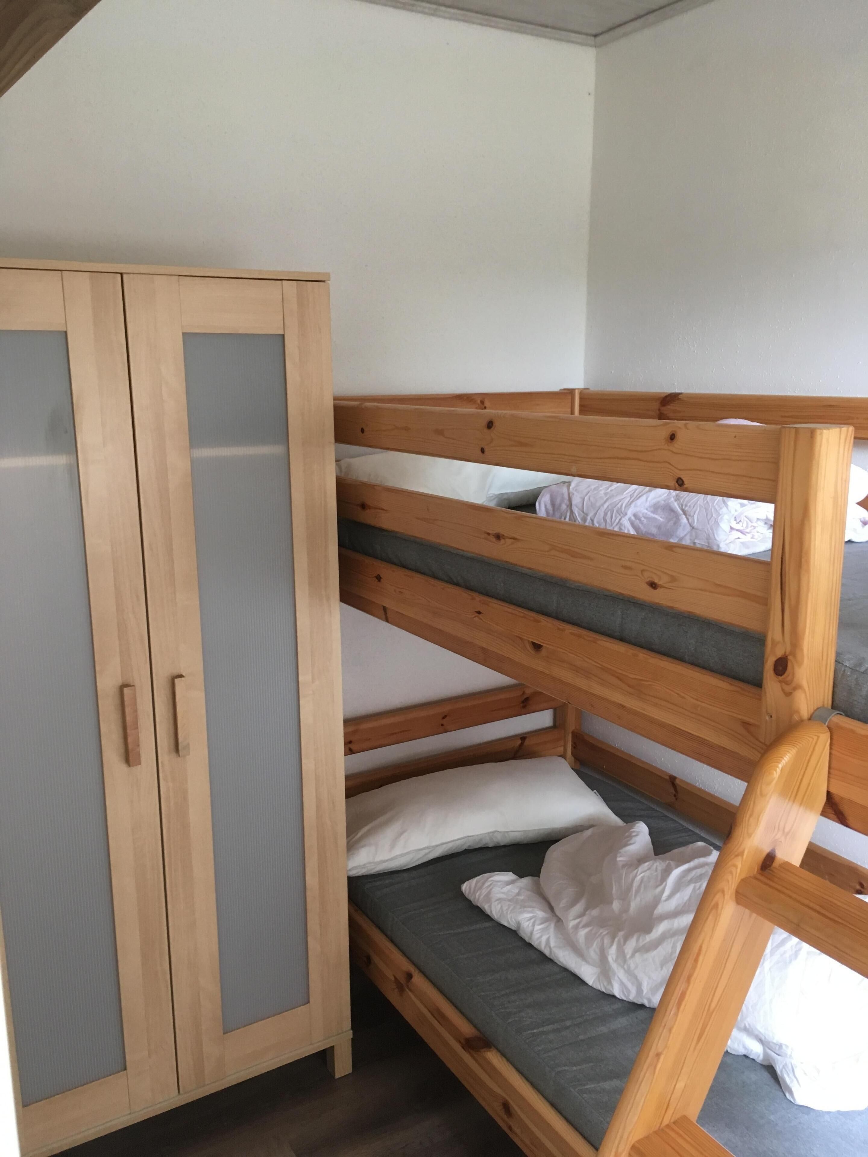 A bunk bed with a wooden frame and grey bedding, a wooden wardrobe with a mirrored door, and a plain white wall.