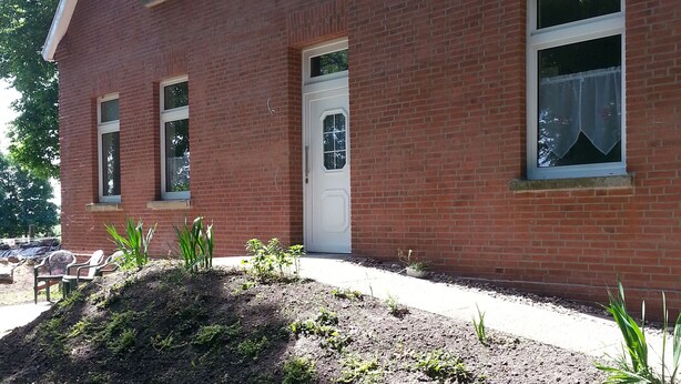 A red brick building with a white door, windows, and a small garden area with plants and a mound of soil. - Holidays with pony rides, WiFi, TV, dishwasher, oven (Butjadingen - Stollhamm)