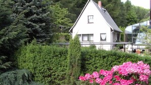 Una casa blanca con techo triangular, rodeada de setos verdes frondosos y un arbusto de flores rosas.