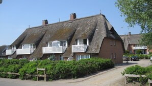 A traditional thatched-roof house with a balcony, surrounded by lush greenery and a gravel driveway. - Seeschlößchen Thatched Cottage, "Westmöwe" Vacation Rental, just 200 meters from the beach (Büsumer Deichhausen)