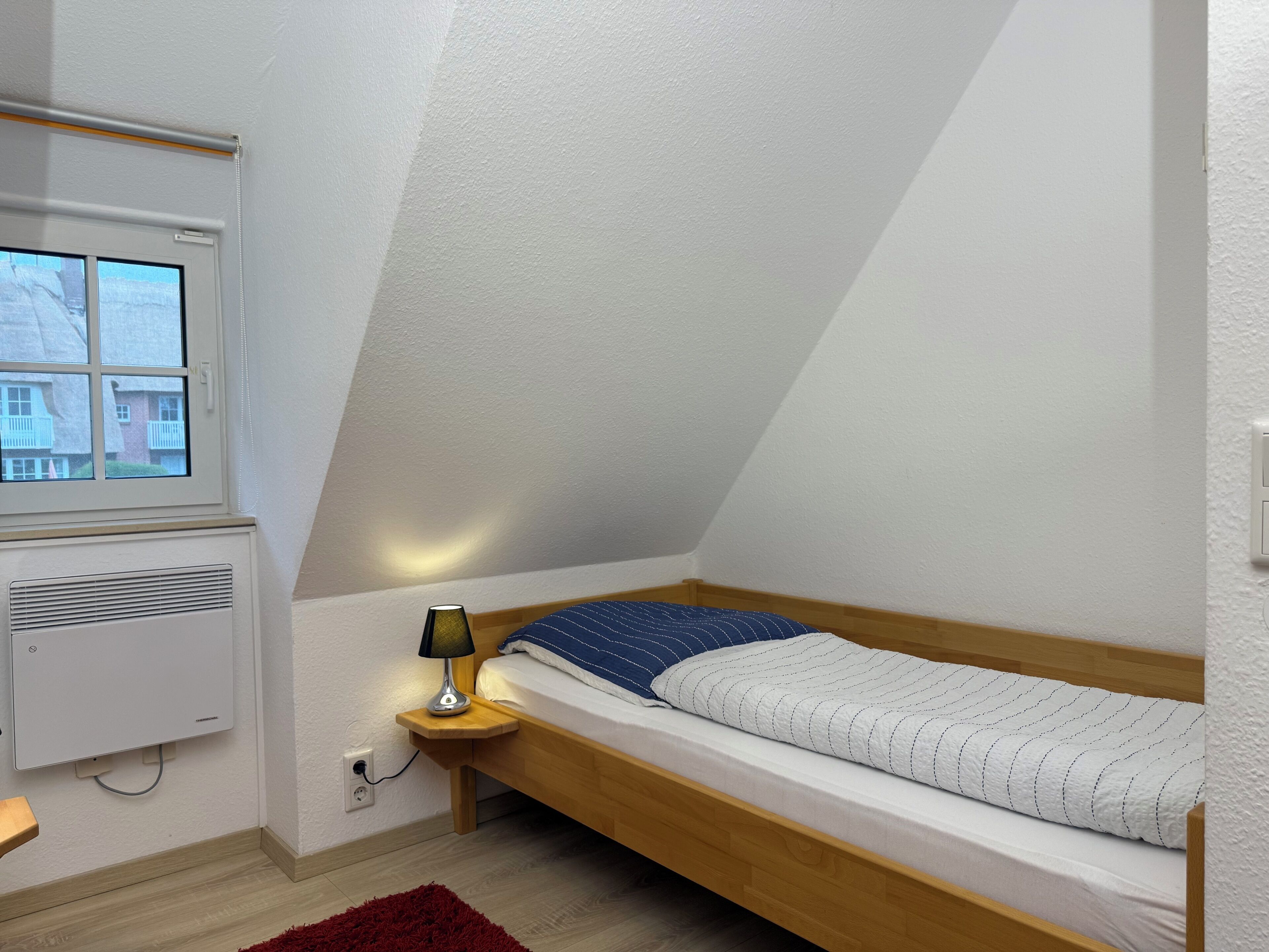 A small, single-bed room with a sloped ceiling, a window, a heater, and a nightstand with a lamp.