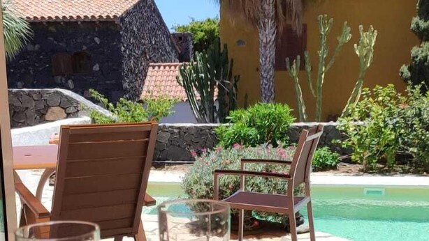 Una mesa con tres vasos de bebidas, una silla de madera y vistas a una piscina y edificios.