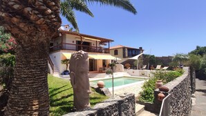 Una casa de dos pisos con piscina, rodeada de follaje y palmeras.