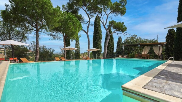 A clear swimming pool with lounge chairs and umbrellas, surrounded by tall trees. - Apartment with a beautiful covered terrace in front of the pool (Torrita di Siena)