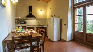 A kitchen with a wooden table, chairs, a refrigerator, and a window with a view of greenery. - Apartment with a beautiful covered terrace in front of the pool (Torrita di Siena)