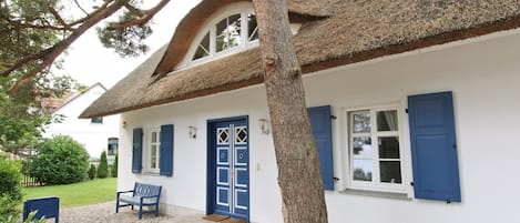 Um edifício branco com telhado de palha, persianas azuis e um banco na parte externa.
