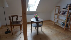 Une pièce avec un plancher en bois, une table ronde ornée d’un vase de fleurs, une chaise en bois, une bibliothèque et une lucarne.