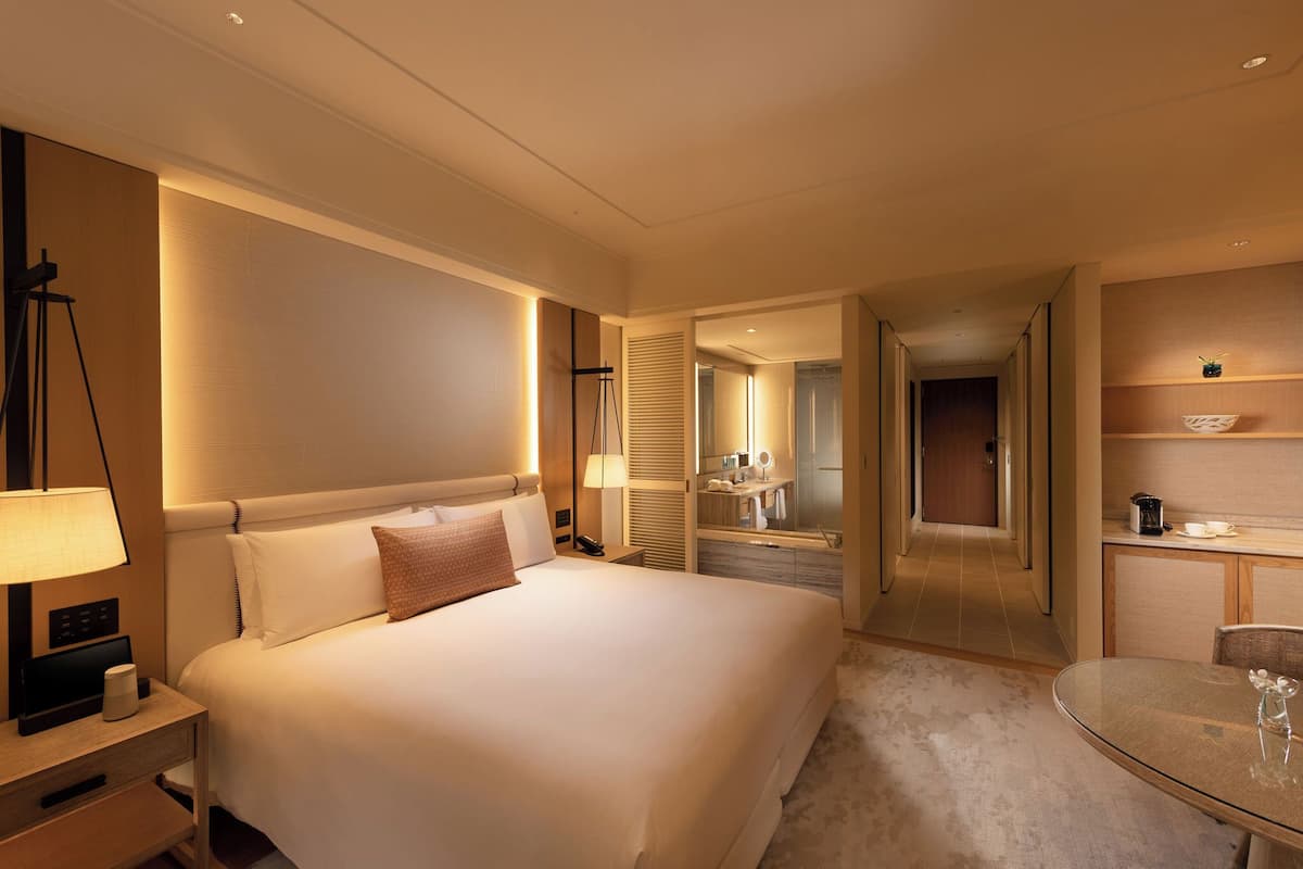 A hotel room with a large bed, bedside table, lamp, and a small table with a glass top.