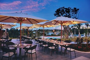 An outdoor dining area with multiple tables and chairs, large umbrellas, and a pool in the background. - Halekulani Okinawa (Onna)