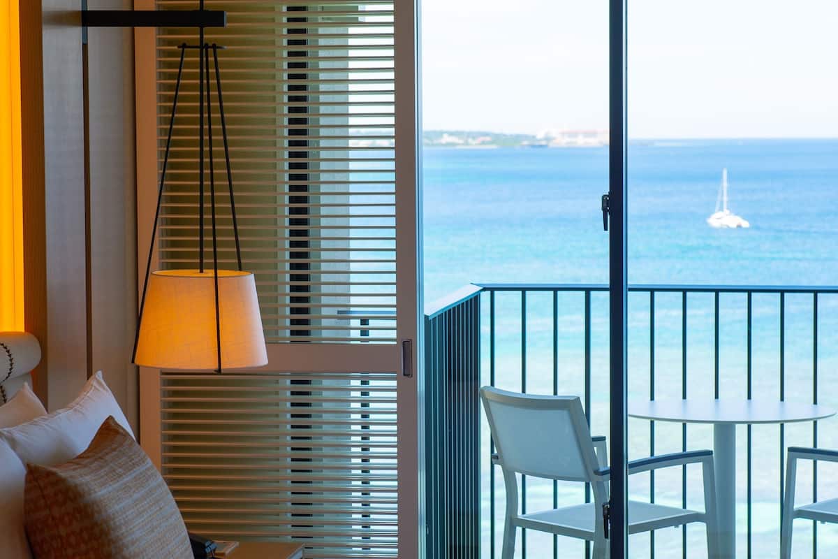 A hotel room with a bed, a table, two chairs, a lamp, and a view of the sea.