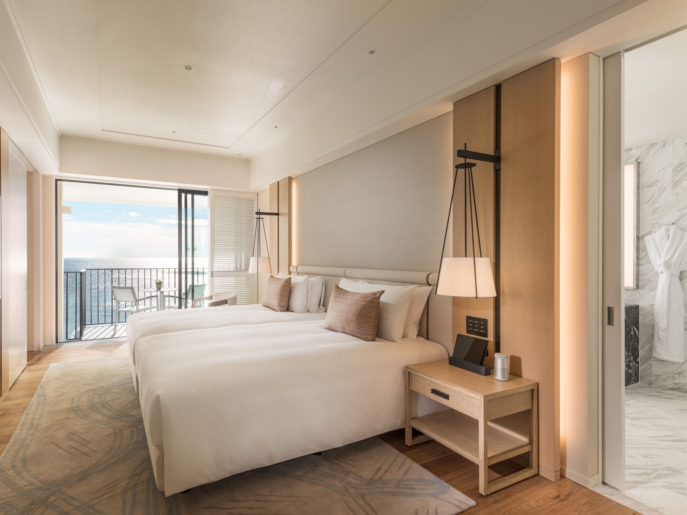 Ein modernes Hotelzimmer mit einem großen Bett, einem Nachttisch mit Lampe und Meerblick von einem Balkon, sowie einer Marmorwand.