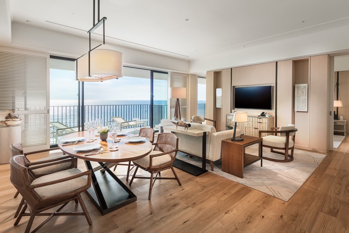 A modern hotel room with a balcony, a dining area with a round table, and a living area with a TV and seating.