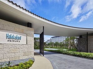 A hotel entrance with a stone facade, a clear sky, and a view of a modern building in the background. - Halekulani Okinawa (Onna)