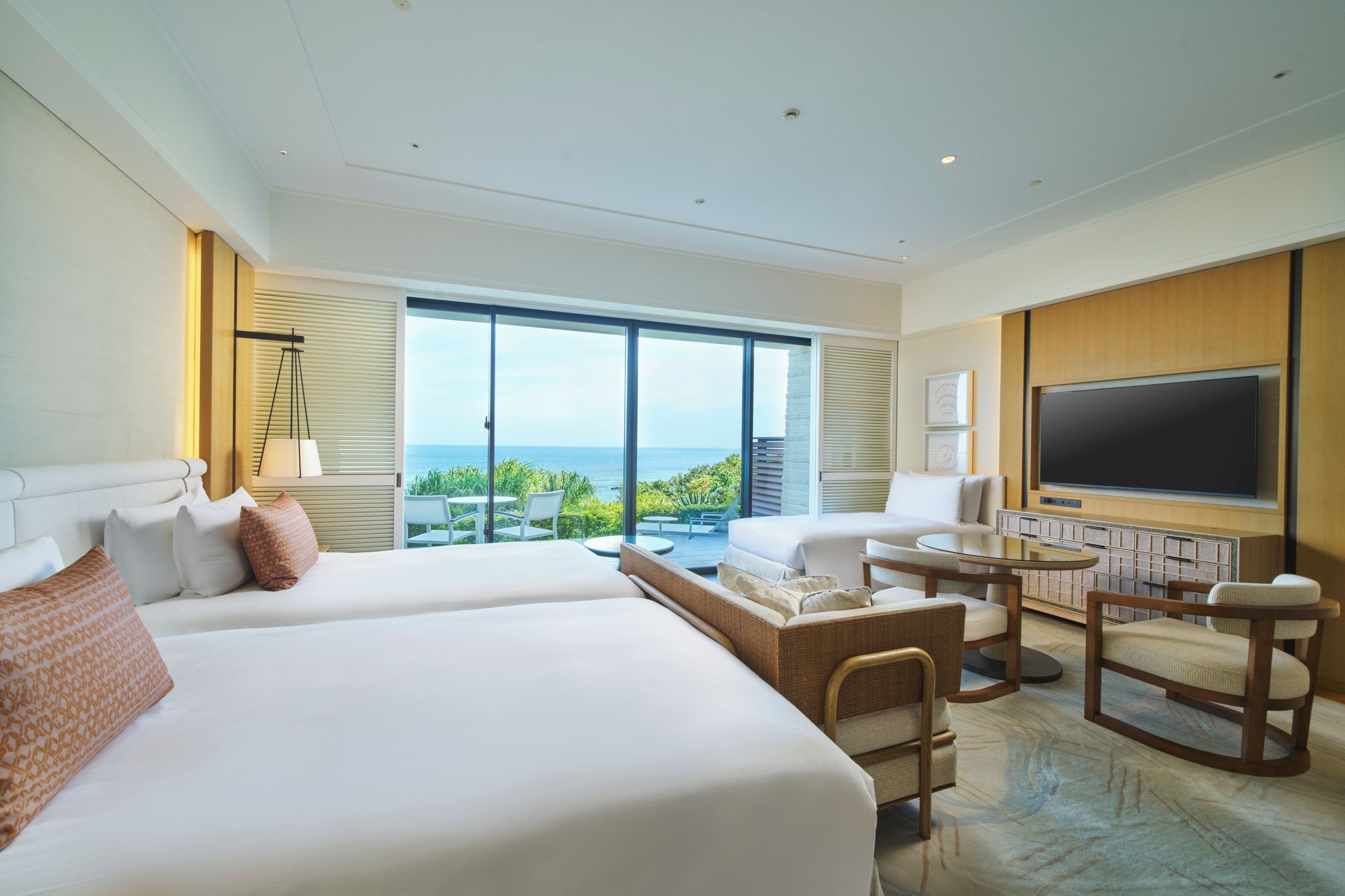 Habitación de hotel con dos camas, televisión, zona de estar y un amplio ventanal con vista al océano.