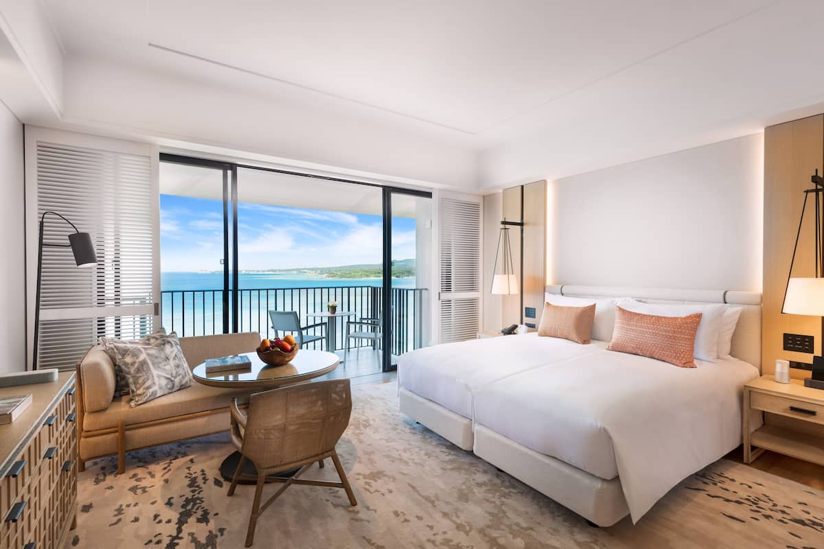 A hotel room with a large bed, a balcony with a view of the sea, a sitting area with a table and chairs, and a desk with a lamp.