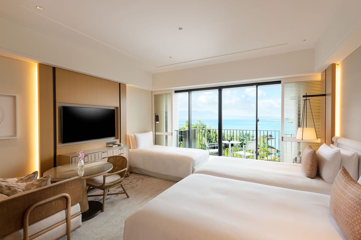 A modern hotel room with a large TV, two beds, a dining area, and a balcony with a view of the sea.