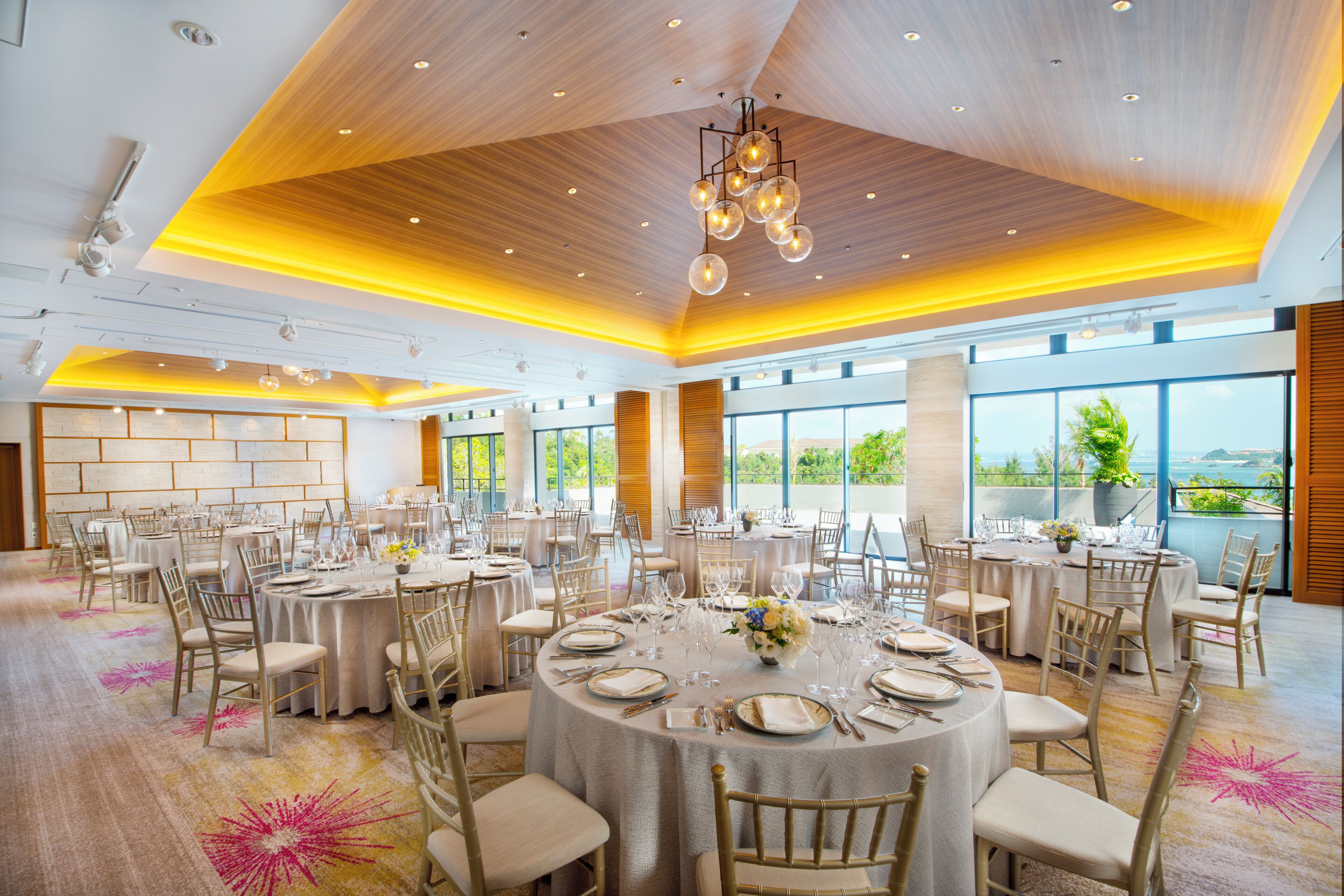 A spacious banquet hall with round tables set for dining, large windows offering views, and a modern ceiling design.