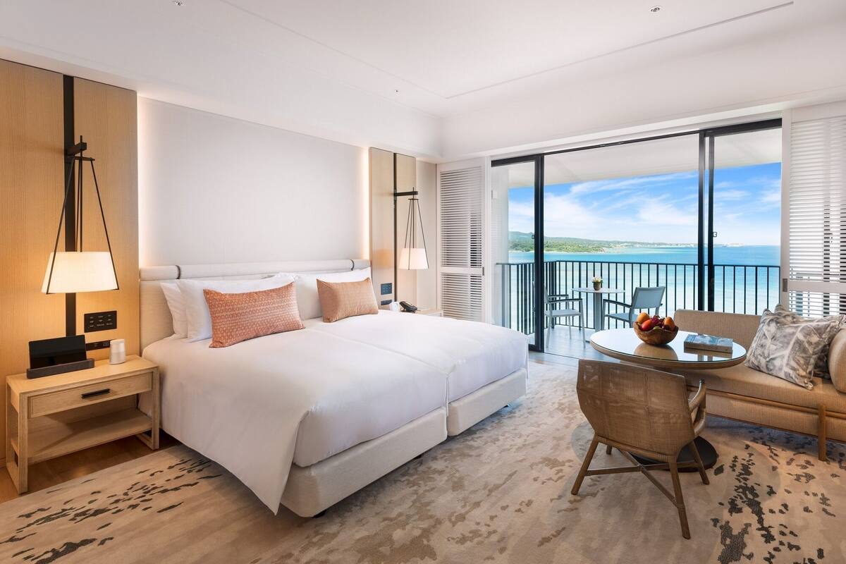 A modern hotel room with a large bed, a desk, a chair, and a balcony with a view of the sea.