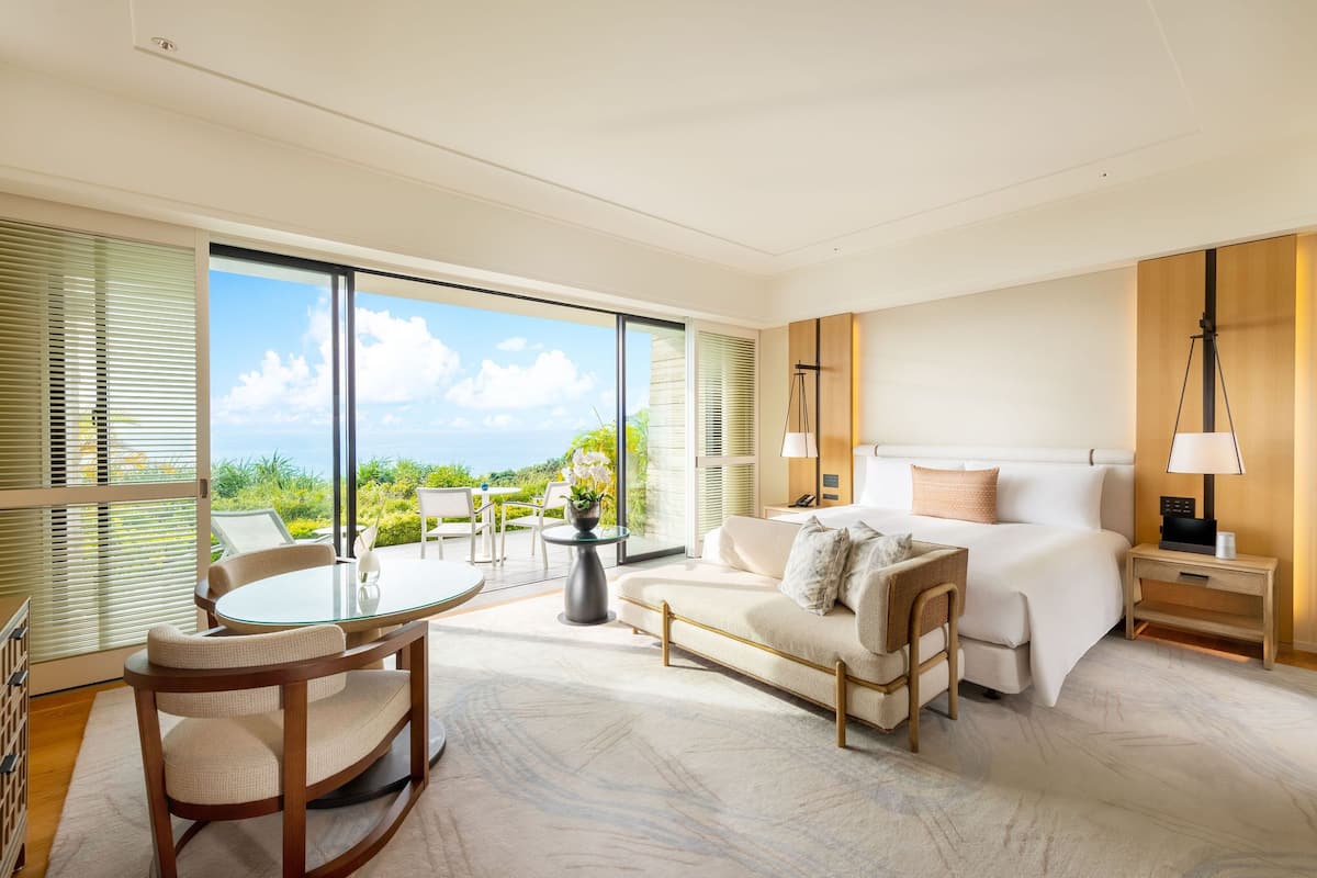 A modern hotel room with a large balcony, a bed, a sitting area with a table and chairs, and a view of the ocean.