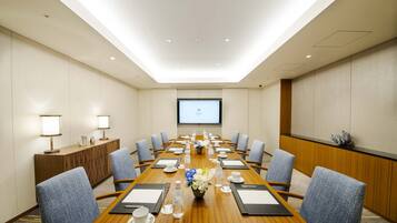 A conference room with a long table, chairs, a projector screen, and a wooden cabinet with a lamp.