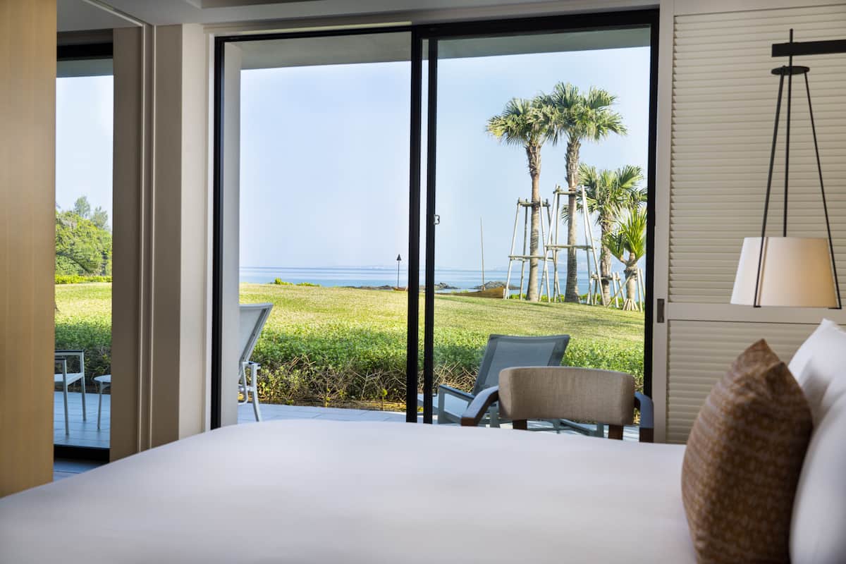 A hotel room with a large bed, a chair, and a view of the beach and palm trees through the sliding glass door.