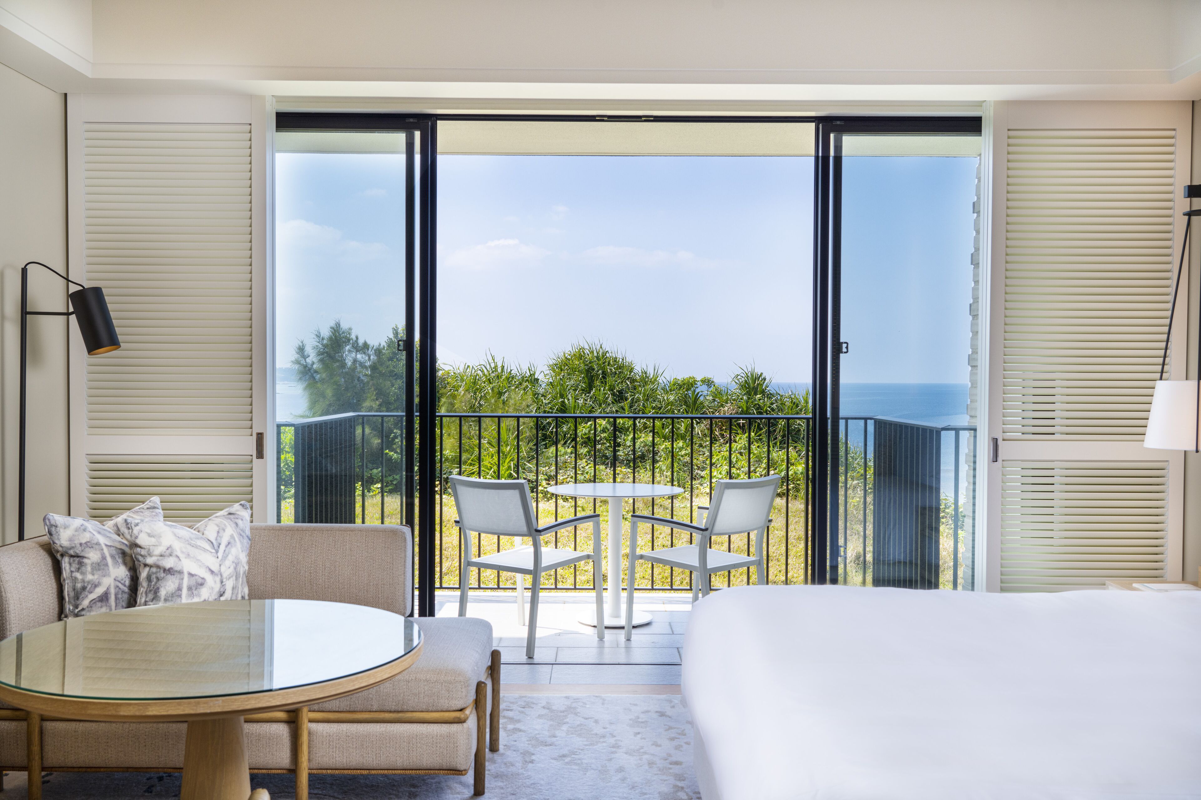A hotel room with a balcony, a round glass table, two chairs, a sofa, and a bed.