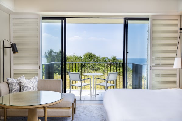 Une chambre d’hôtel avec un balcon, une table ronde en verre, deux chaises, un canapé et un lit.