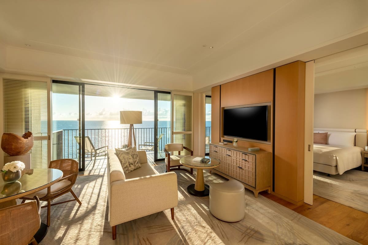 A spacious living room with a large TV, comfortable seating, a dining area, and a balcony with a view of the sea.