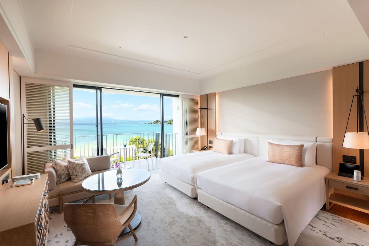 A hotel room with two beds, a sofa, a small table, and a balcony with a view of the sea.