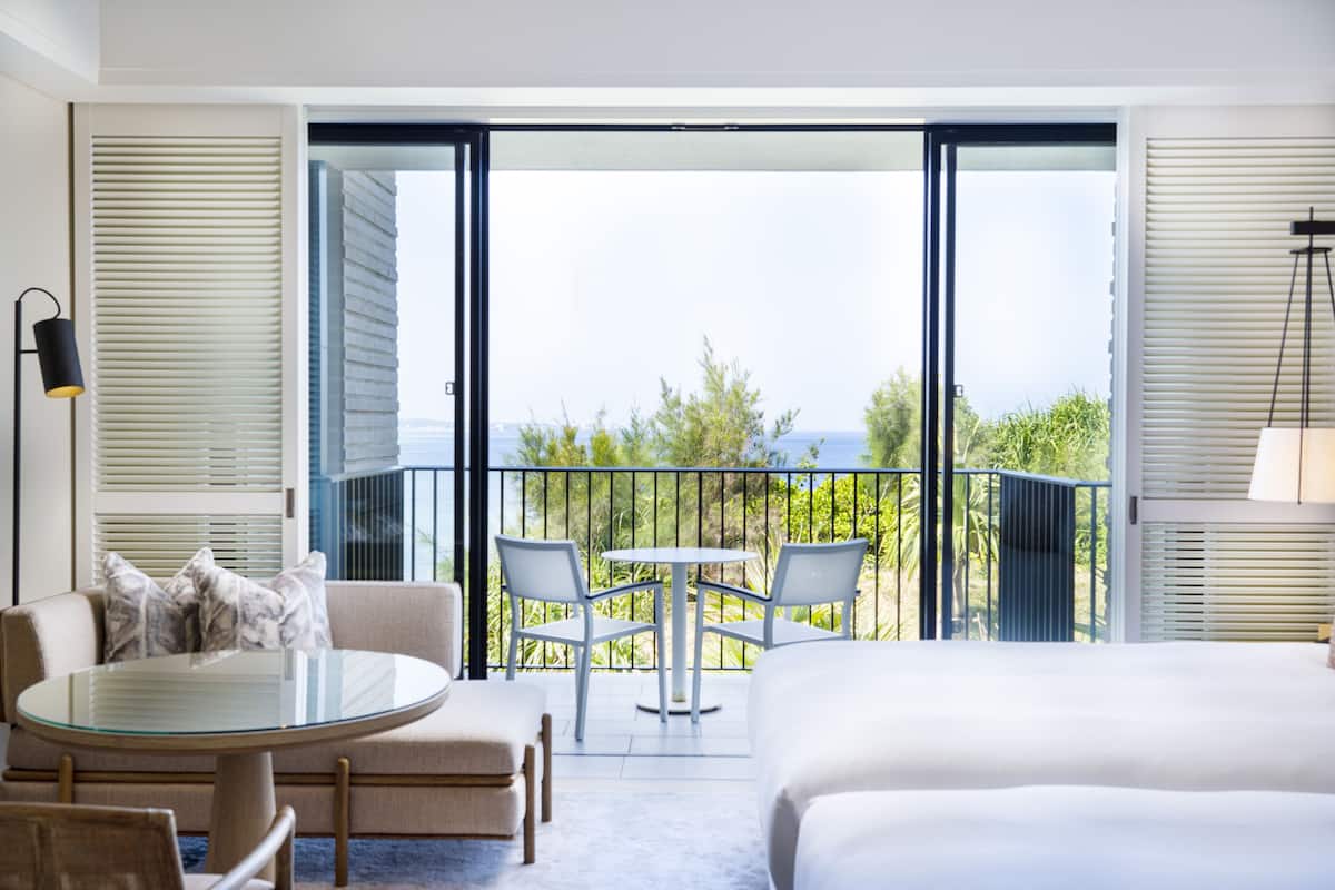A hotel room with a balcony, a bed, a round glass table, two chairs, and a sofa.