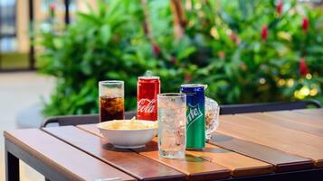 A wooden table with a bowl of chips, two glasses of soda, and two cans of soda.
