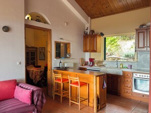 A compact kitchen with a wooden island, a dining area with a red sofa, and a view of greenery outside. - Country house in the olive grove with breathtaking views for every season (Messinia)