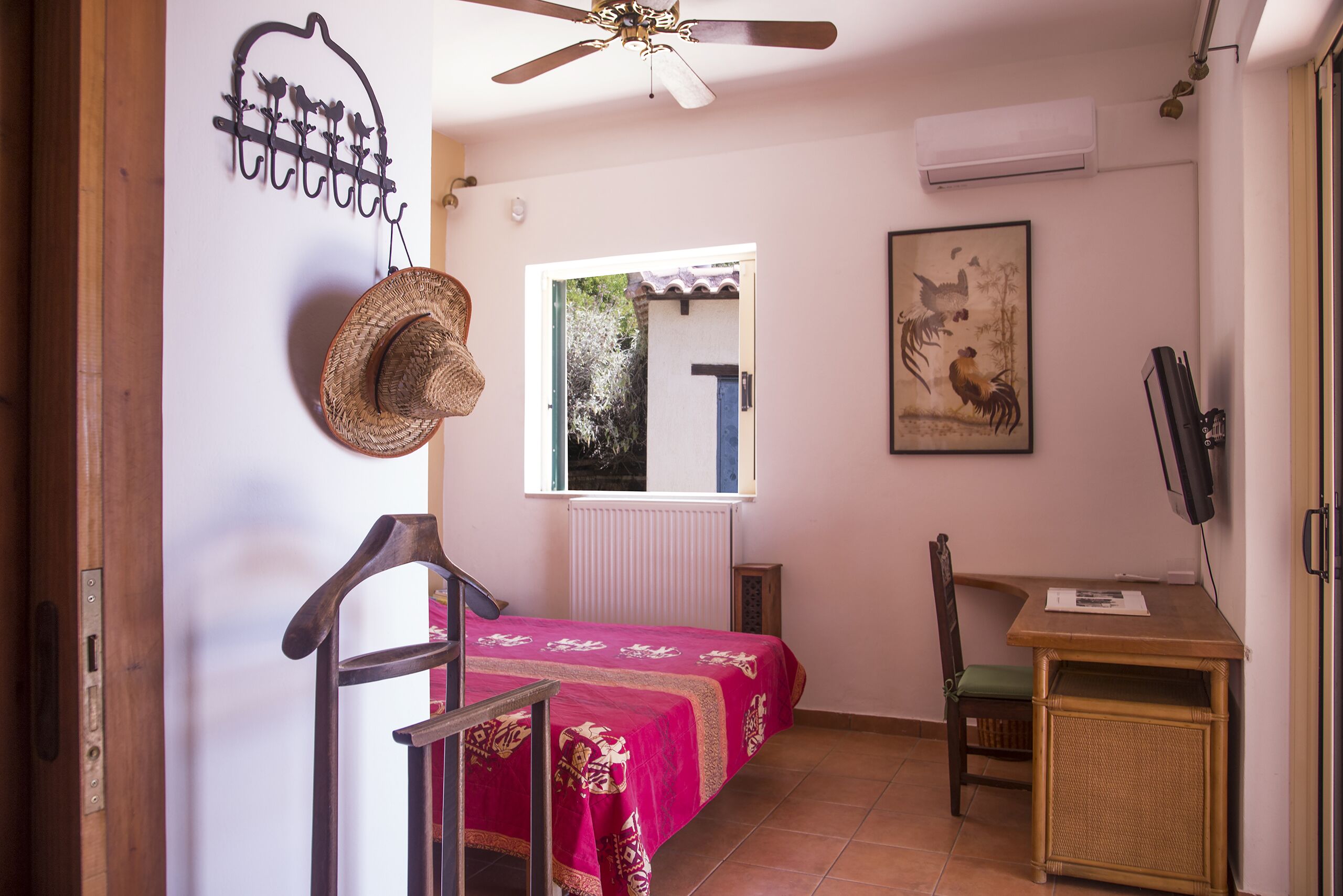 Ein Schlafzimmer mit Bett, Sessel, Nachttisch, Deckenventilator, Fenster und Wandbild.