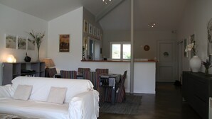 Een ruime woonkamer met een witte bank, een eettafel met gestreepte stoelen en een keuken op de achtergrond.